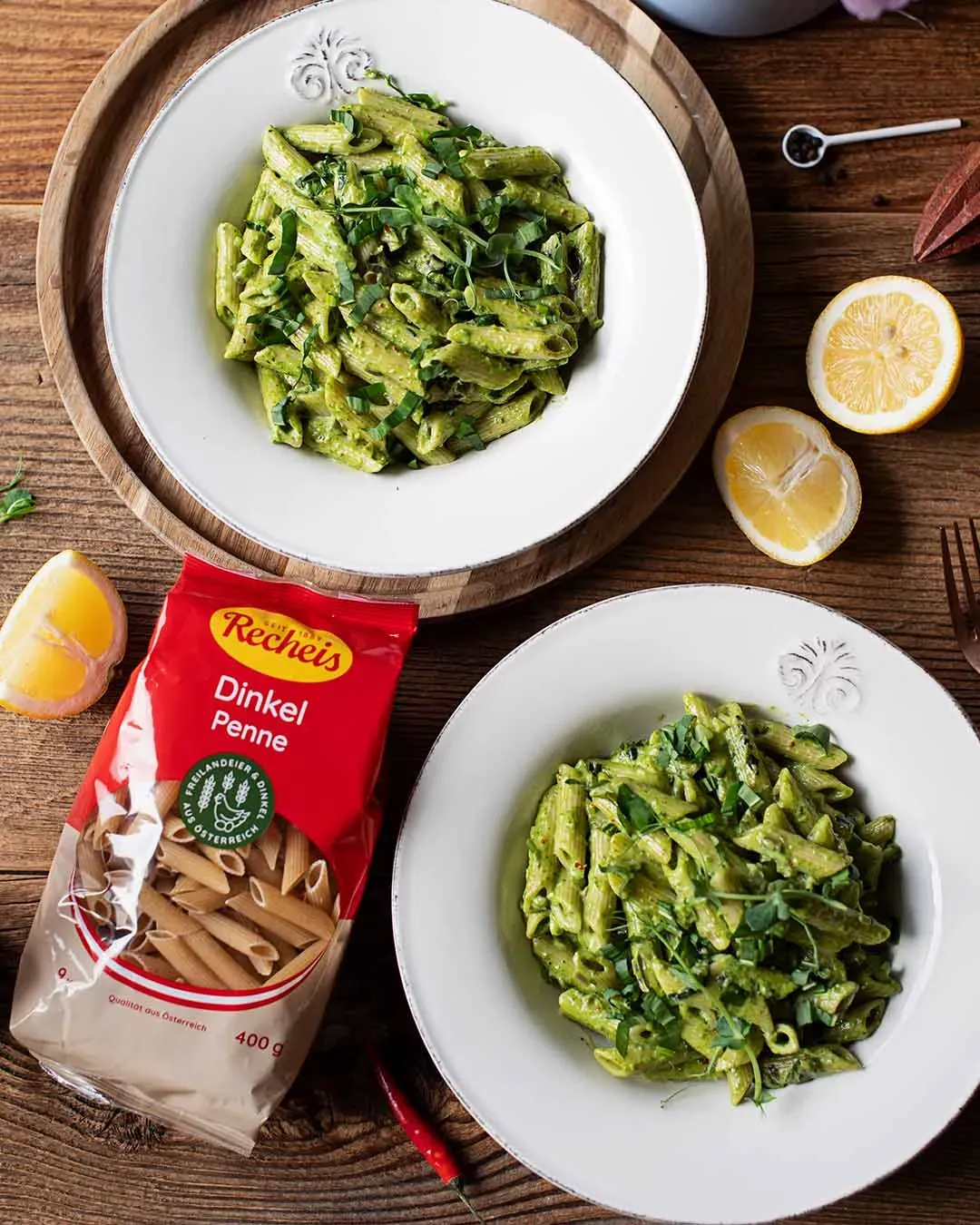 Nudelpfanne mit Bärlauch und Dinkel Penne auf zwei Tellern neben der Packung Dinkel Penne auf Holztisch