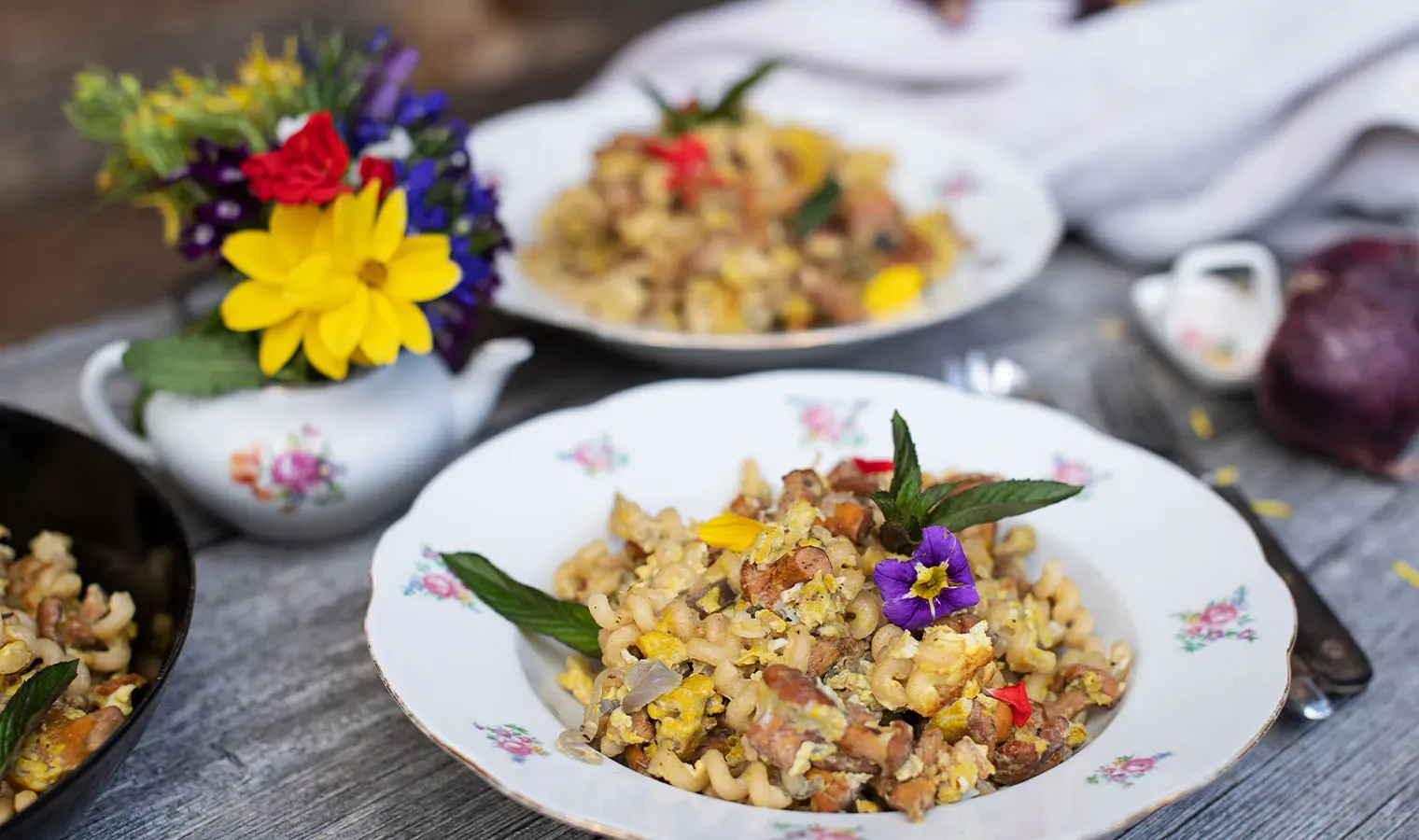 Nudelpfanne mit Eierschwammerl und bunten essbaren Blüten auf einem Teller auf Holztisch mit Blumenstrauß