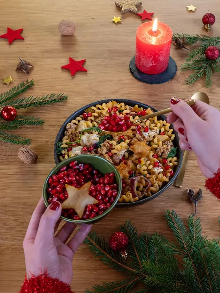 Person bestreut weihnachtliche Nudelpfanne mit Granatapfelkernen vor Weihnachtsdekoration