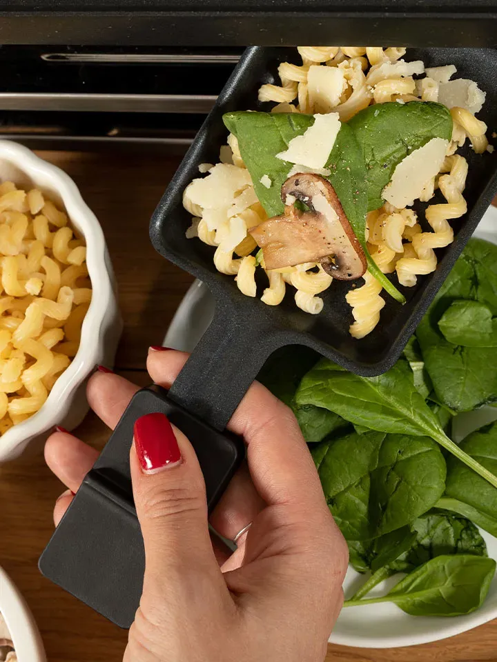 Hand hält eine Pfanne mit Nudelraclette aus Pasta, Spinatblatt, Pilz und Käse