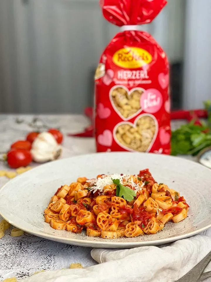 Herzförmige Pasta in Tomatensauce mit Speck und Pfefferoni und geriebenem Käse auf Teller vor Nudelpackung