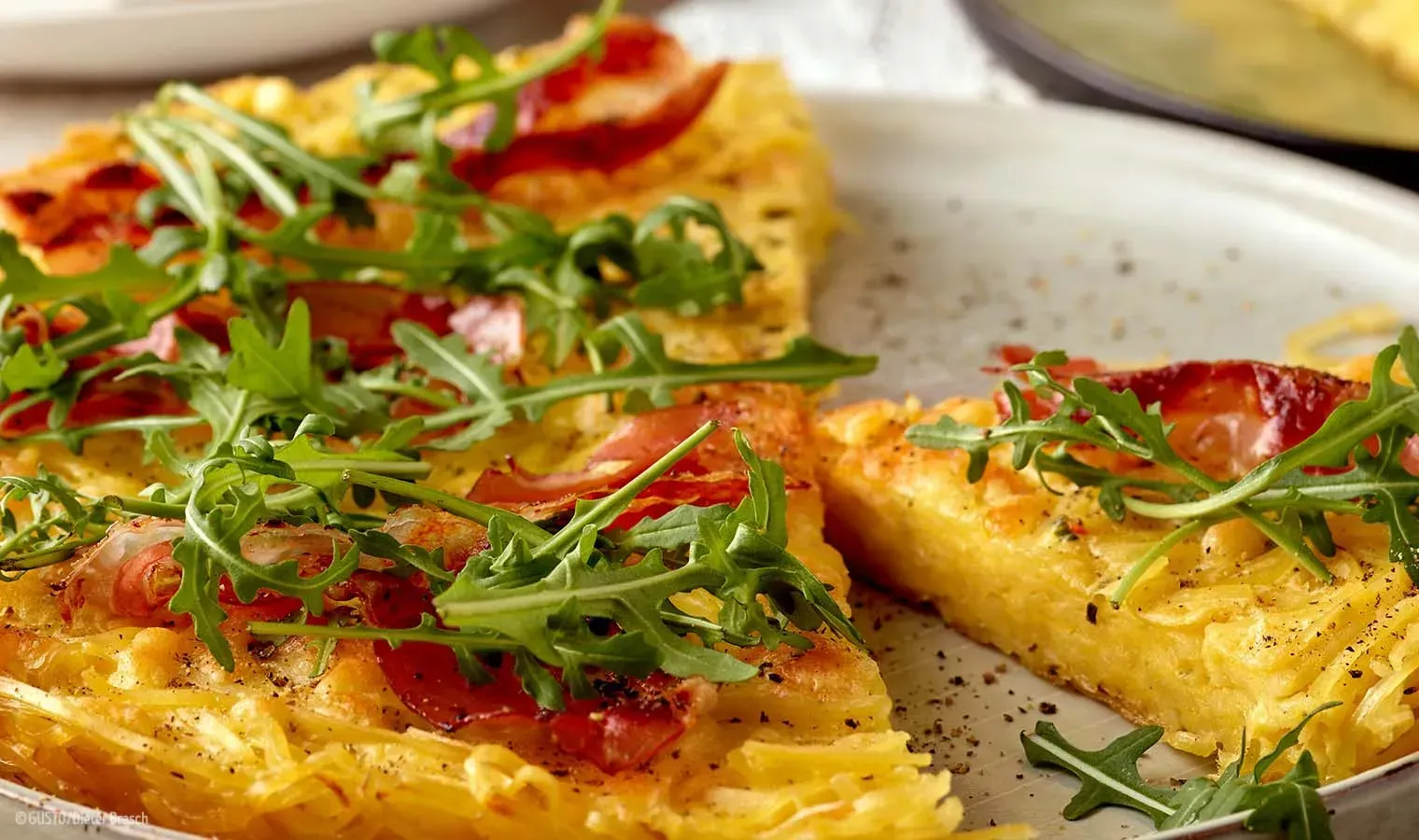 Nahaufnahme der Nudelrösti mit knusprigem Speck, Rucola und grob gemahlenem Pfeffer