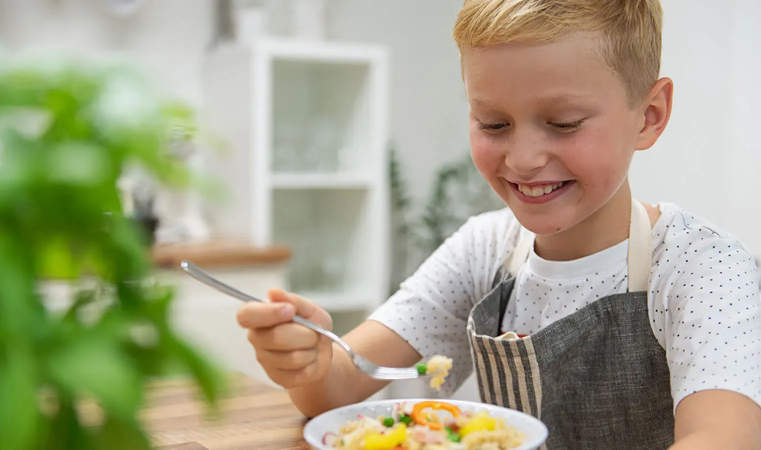 Lächelnder Junge isst in der Küche mit einer Gabel einen Teller Nudelsalat