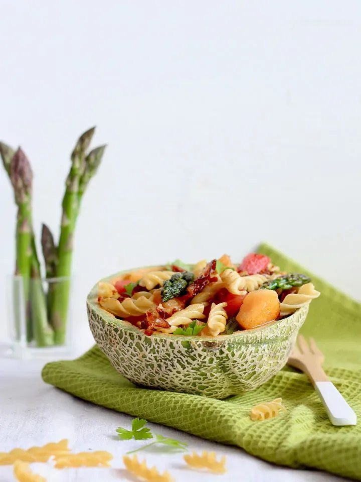 Low-Carb-Nudelsalat mit Speck und Spargel in einer Melonenschale als Schüssel auf grünem Tuch