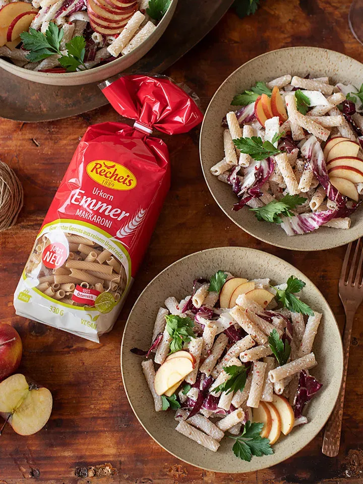 Nudelsalat mit Apfel und Radicchio auf zwei Tellern, daneben eine Packung Emmer-Makkaroni auf Holztisch
