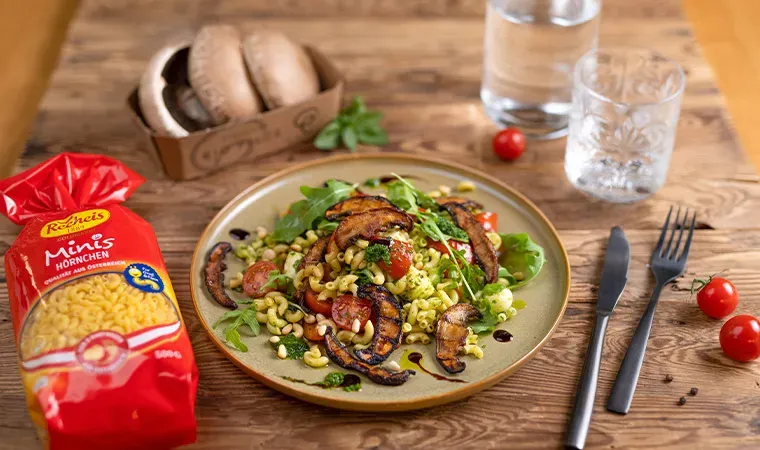 Nudelsalat mit Portobello-Bacon, gebratenen Pilzen und Tomaten auf Holztisch mit Besteck und Wasser