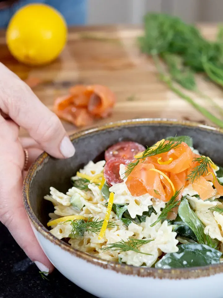 Hand hält eine Schüssel Nudelsalat mit Räucherlachs und Dill auf Holztisch