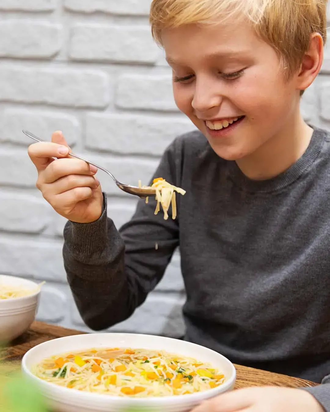 Lachender Bub isst Gemüsesuppe mit Dinkel Suppennudeln an einem Holztisch