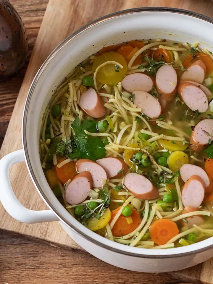 Nudelsuppe mit Würstchen und Gemüse im Topf auf einem Holzbrett