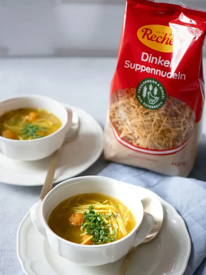Zwei Suppentassen mit Nudelsuppe und Karottenknöderln, dahinter die Packung Dinkel Suppennudeln
