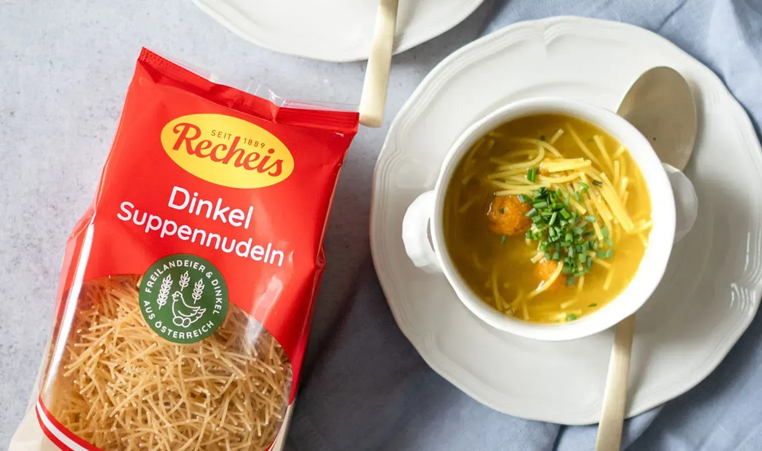 Aufsicht auf eine Tasse Nudelsuppe mit Karottenknöderl und Schnittlauch neben einer Packung Recheis Dinkel Suppennudeln