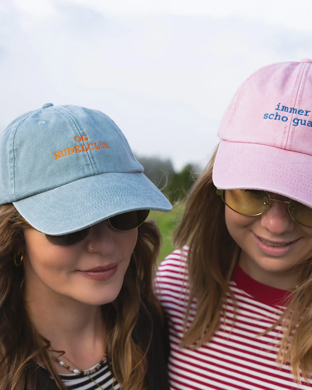 Zwei Frauen mit Baseballcaps und Sonnenbrillen lächeln nach unten