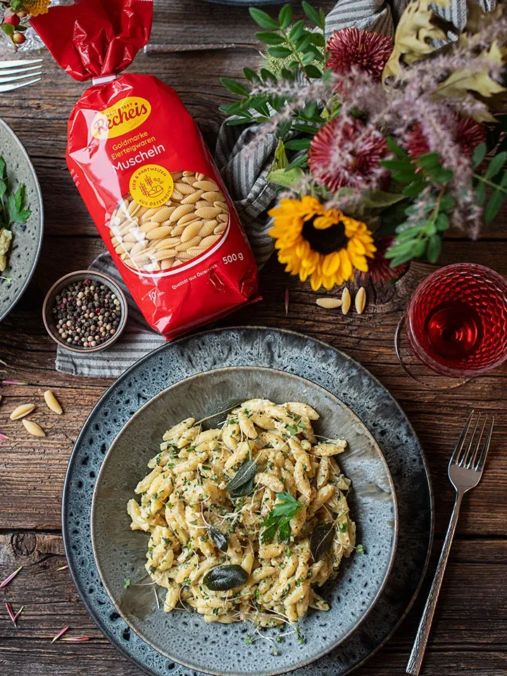 One-Pot-Nudelpfanne mit Kräutern und Muschelnudeln auf einem Teller neben roter Verpackung und Blumen auf Holztisch
