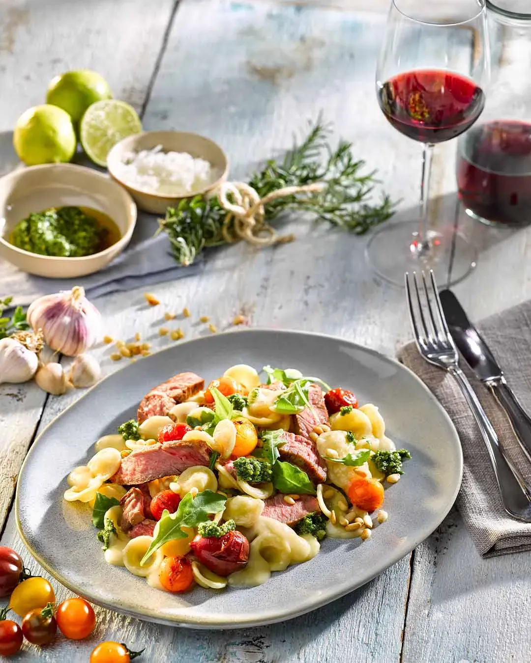 Orecchiette Tagliata di Manzo mit Steakstreifen, Tomaten und grünem Pesto, daneben Rotweinglas und Kräuter