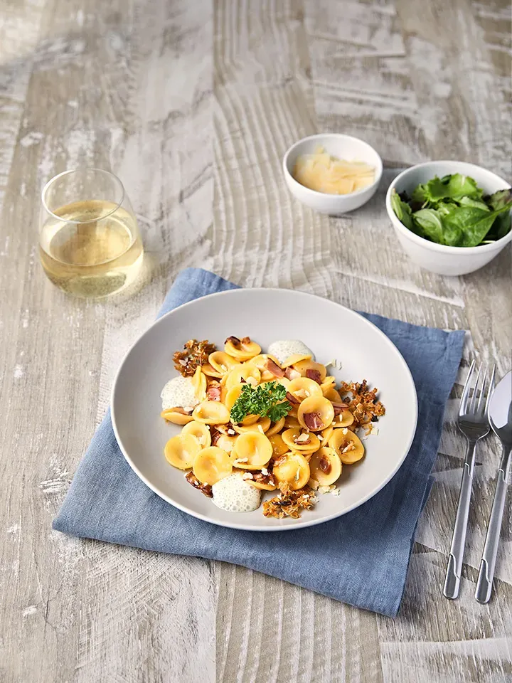 Orecchiette Carbonara serviert mit Salat, Käse und Glas Weißwein auf Holztisch