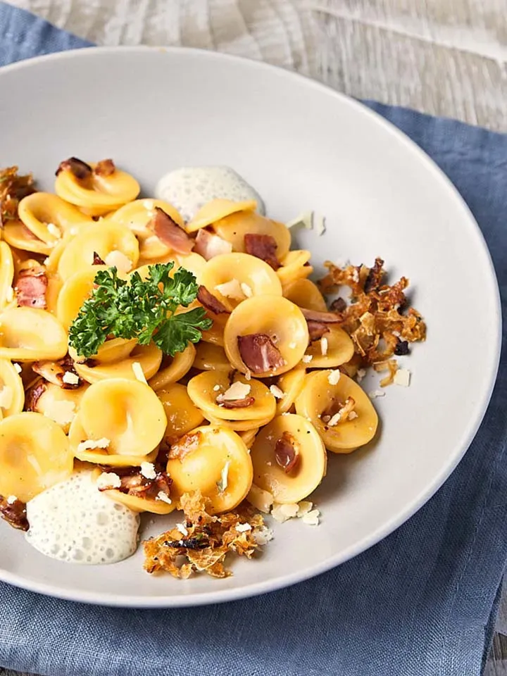 Orecchiette Carbonara mit Speck, Kräutern und Zwiebelespuma auf blauem Tuch