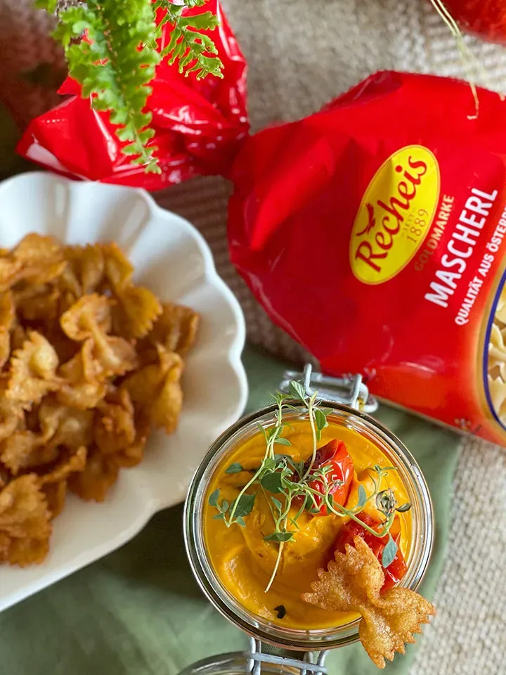 Paprika-Nudelchips mit Kürbisdip: Glas mit Dip, gebratene Farfalle und rote Nudelpackung
