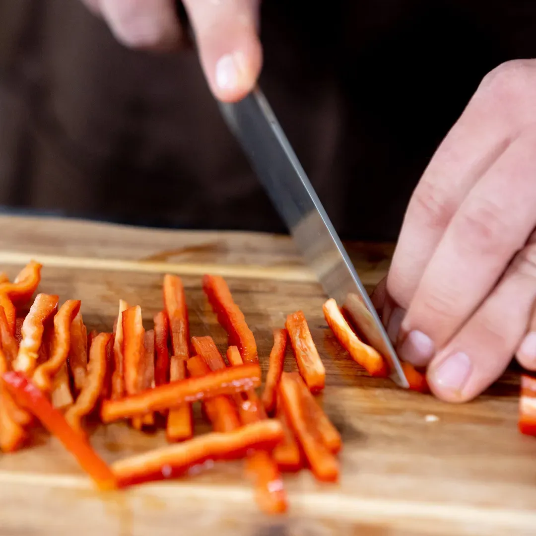 Hände schneiden Spitzpaprika für Pasta Arrabbiata in dünne Streifen auf einem Holzbrett