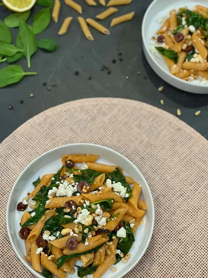 Pasta mit Harissa: Penne mit Spinat, Oliven, Feta und Pinienkernen auf einem Teller auf Tischdecke