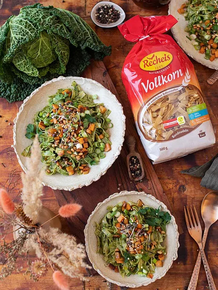Veganes Grünkohlpesto mit Vollkornnudeln auf zwei Tellern, dazu Wirsingkohl und eine Packung Vollkornnudeln auf Holztisch