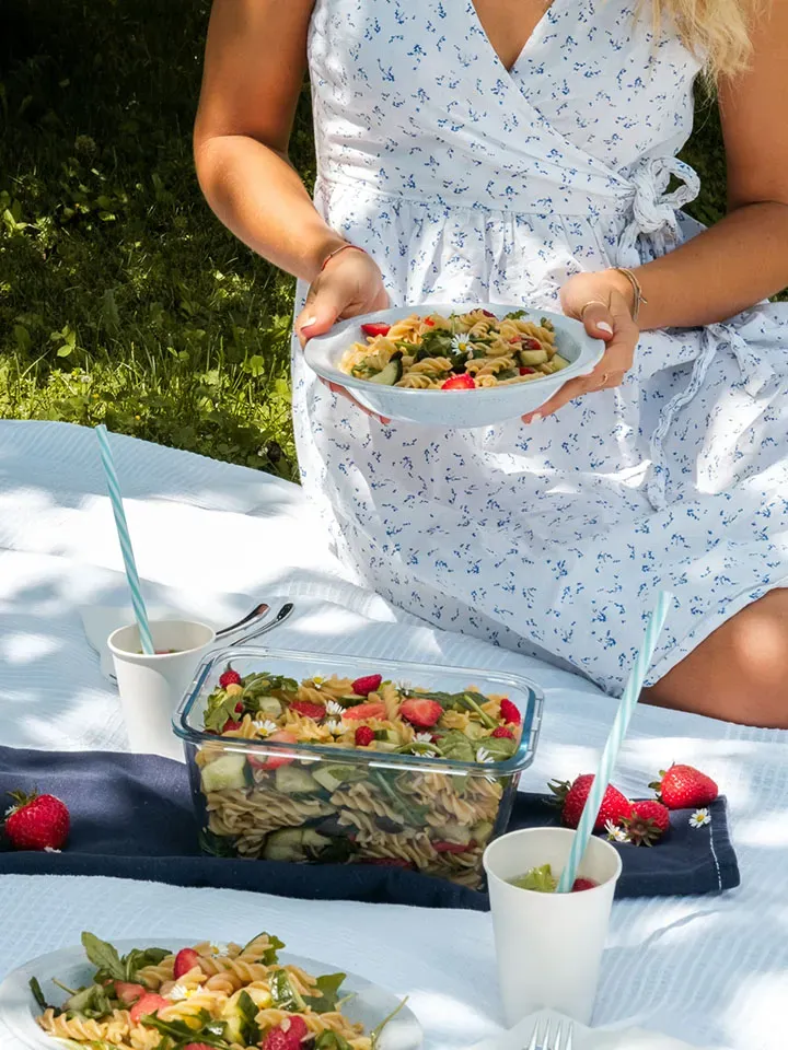 Frau im weißen Kleid hält beim Picknick auf einer Decke im Grünen einen Teller mit Nudelsalat