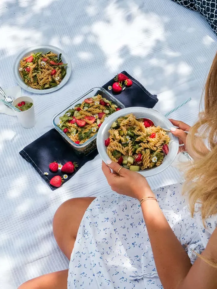 Frau hält beim Picknick auf weißer Decke einen Teller mit Nudelsalat und Erdbeeren