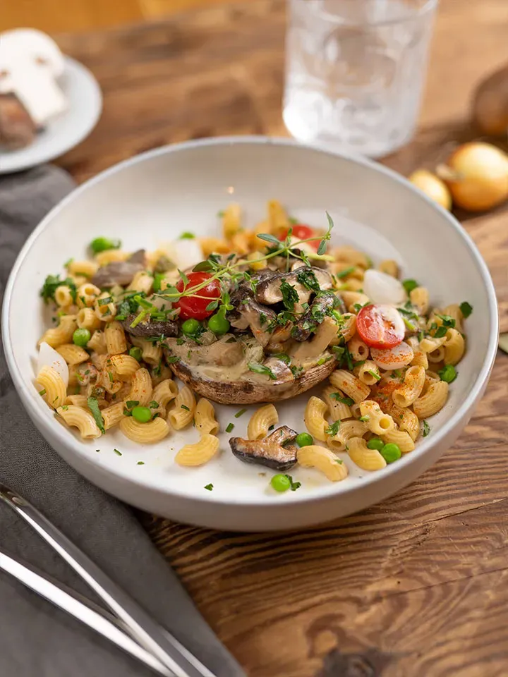 Vegane Pilzpfanne mit Pasta, Pilzen, Erbsen und Kirschtomaten auf einem Teller auf Holztisch