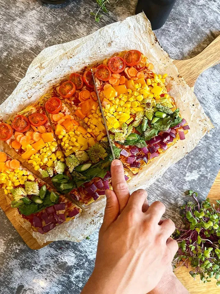 Hand schneidet bunte Gemüsepizza aus Nudeln auf Backpapier auf einem Holzbrett