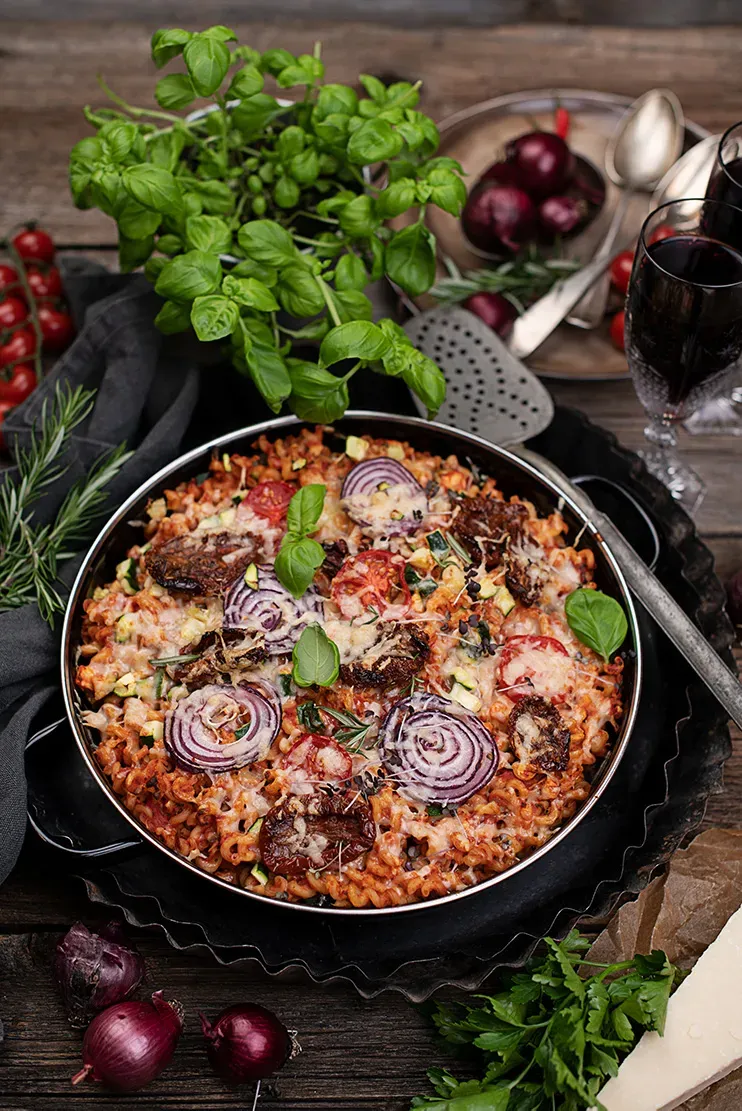 Pizza-Nudel-Auflauf mit Tomaten, Zwiebeln und Käse, umgeben von frischen Kräutern