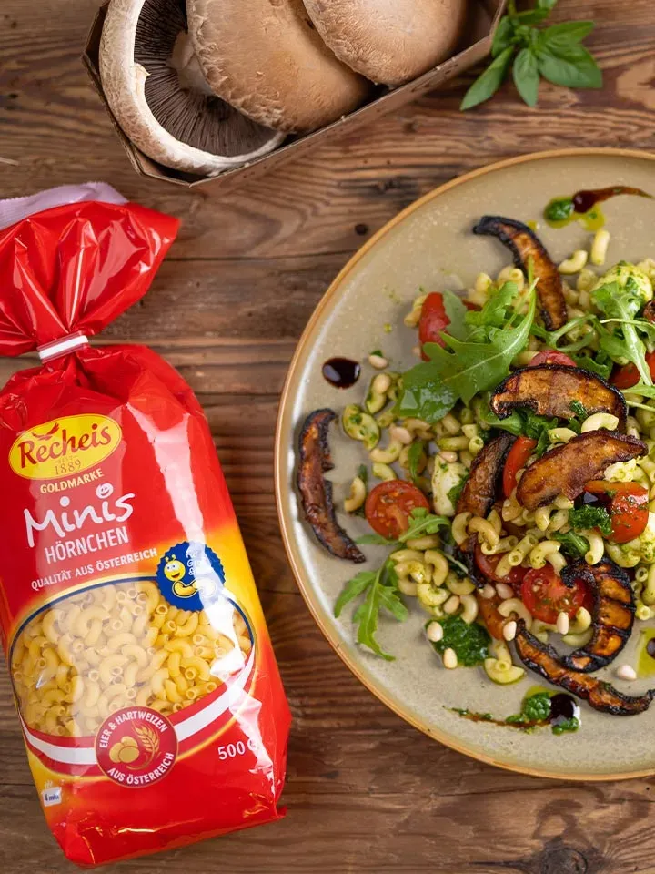 Nudelsalat mit Portobello-Bacon: Teller mit Pasta, gegrillten Pilzen und Tomaten neben Packung und frischen Pilzen