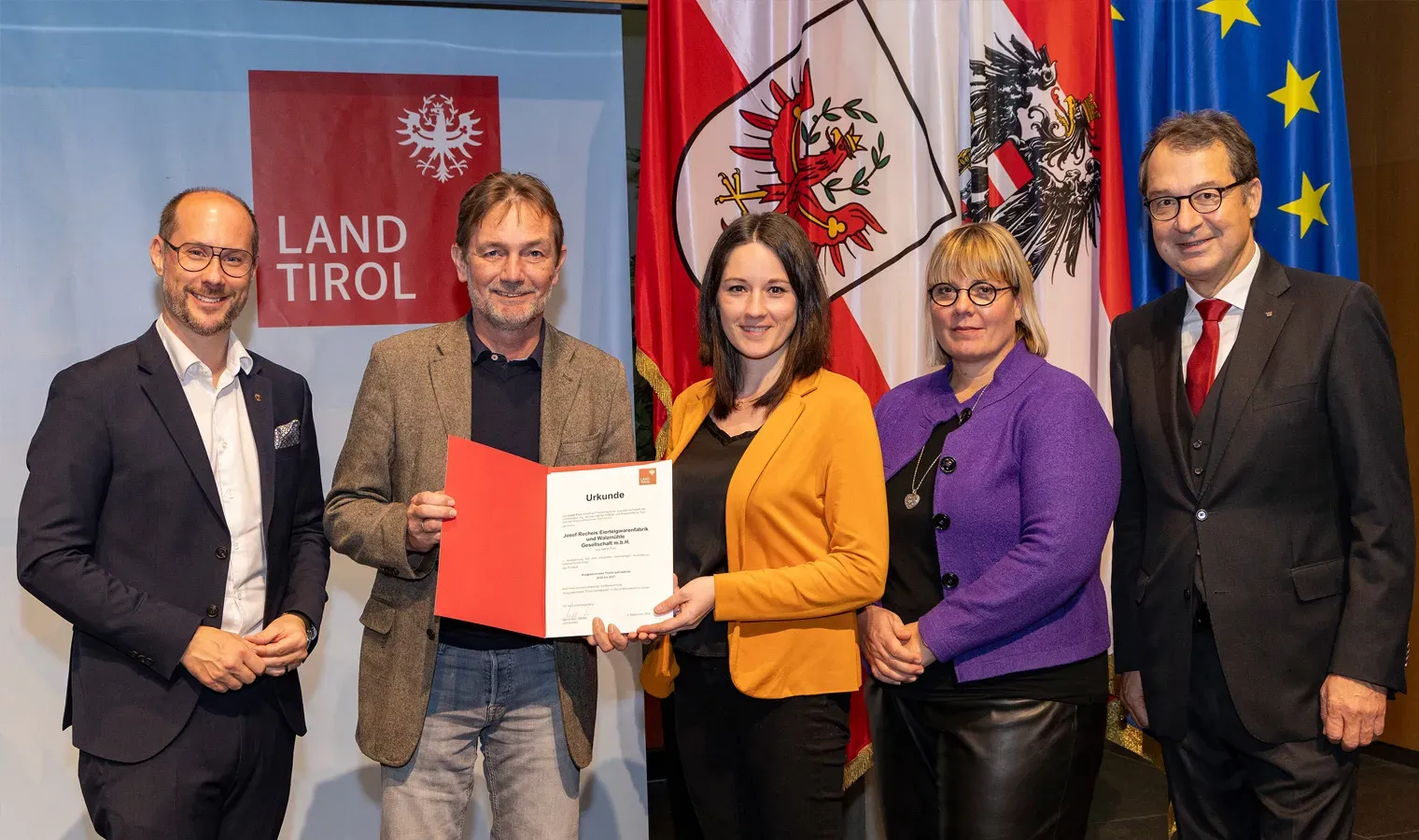 Fünf Personen bei einer Urkundenübergabe zum ausgezeichneten Tiroler Lehrbetrieb vor Tirol- und Europaflaggen