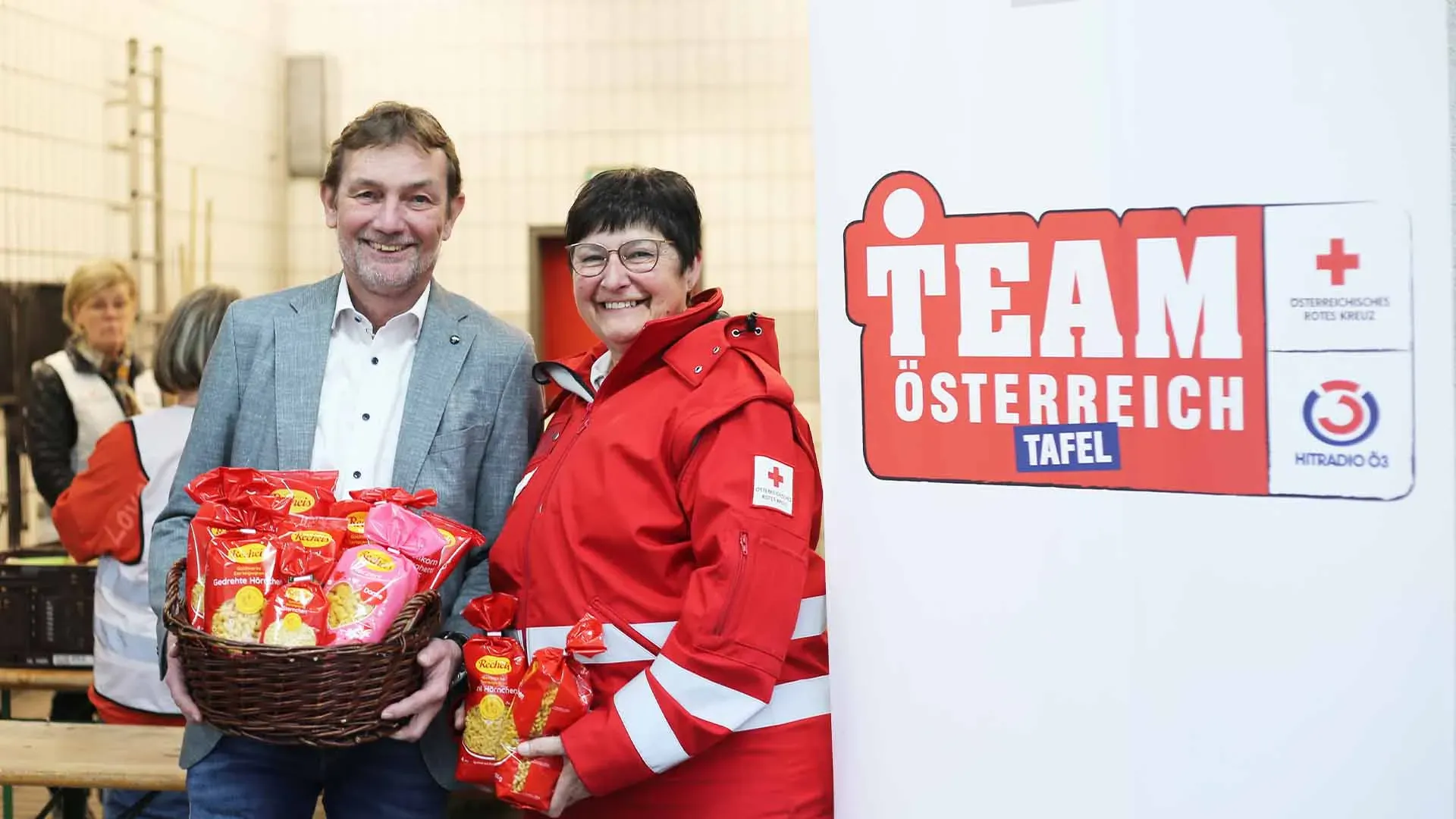 Zwei Personen mit Lebensmitteln vor einem Banner der Team Österreich Tafel, Übergabe 2025