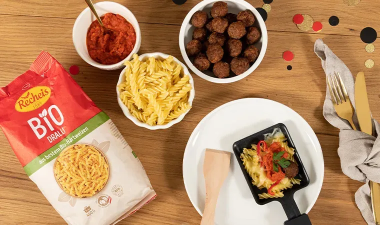 Raclette mit Fleischbällchen, Pasta und Tomatensauce auf einem Teller, daneben Bio-Drahtnudeln auf Holztisch