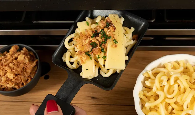 Nudelraclette im Spätzle-Stil: Person hält ein Pfännchen mit Käse, Nudeln und Zwiebelringen