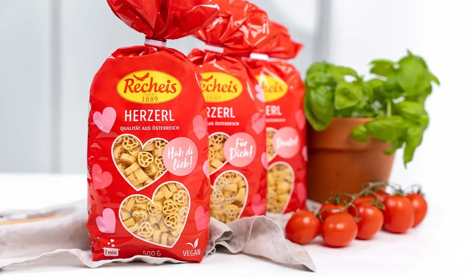 Drei Packungen Recheis Herzerl Nudeln in Herzform, vegan, neben Tomaten und Basilikum auf weißem Untergrund