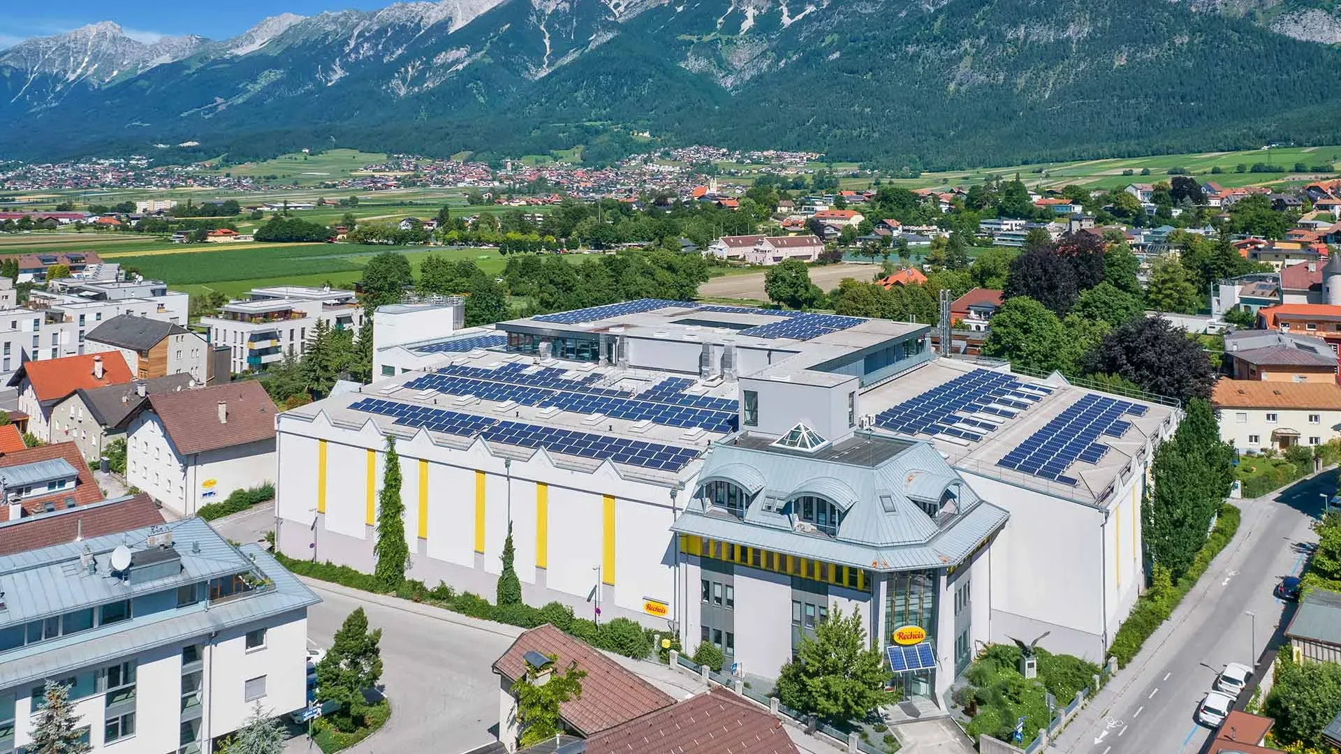 Klimaneutrales Recheis-Gebäude mit Solarpaneelen auf dem Dach vor Bergkulisse und Wohnhäusern