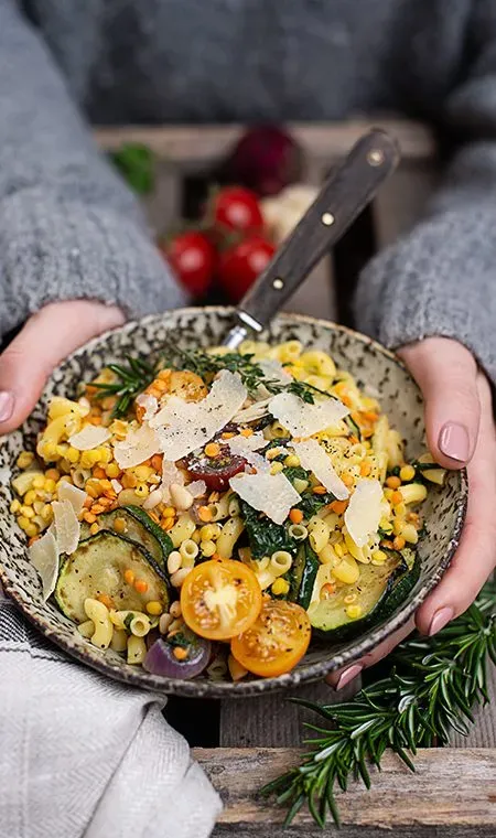 Hände halten eine Schüssel mit glutenfreien Nudeln, Gemüse, Linsen und Parmesanspänen