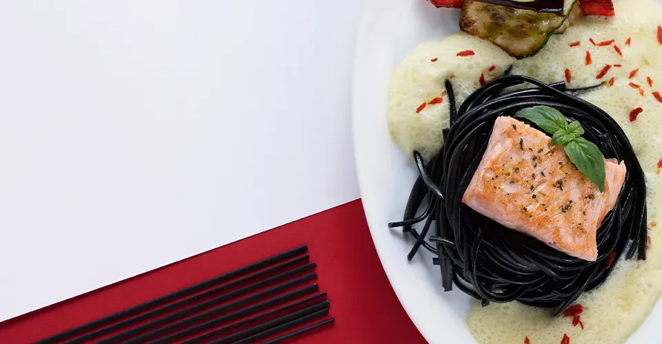 Nahaufnahme: gegrilltes Lachsfilet auf Linguine nero mit Safransauce und Grillgemüse auf weißem Teller