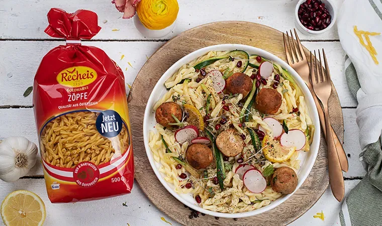 Nudel-Zucchinipfanne mit Lachsbällchen und Gemüse auf Teller neben Packung Recheis Goldmarke Zöpfe