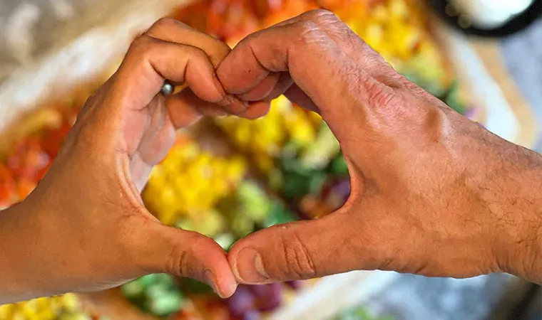 Regenbogen-Pizza aus Nudeln: zwei Hände formen ein Herz vor buntem, unscharfem Hintergrund