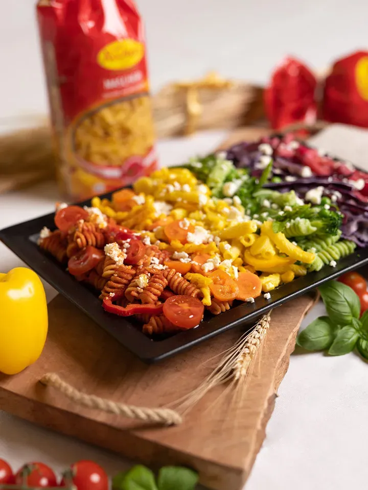 Bunte Regenbogennudeln mit Gemüse und Feta auf einem schwarzen Teller auf Holzbrett