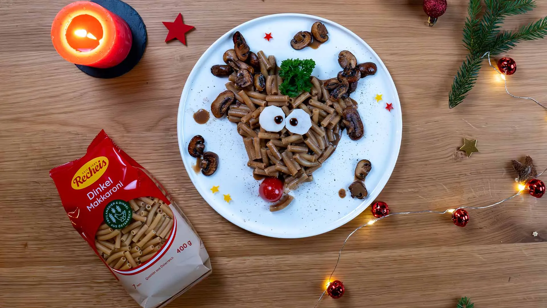 Rentiergesicht aus Dinkel-Makkaroni mit Champignons, Rettich-Augen und Kirschtomaten-Nase, daneben eine Packung Recheis Dinkel Makkaroni