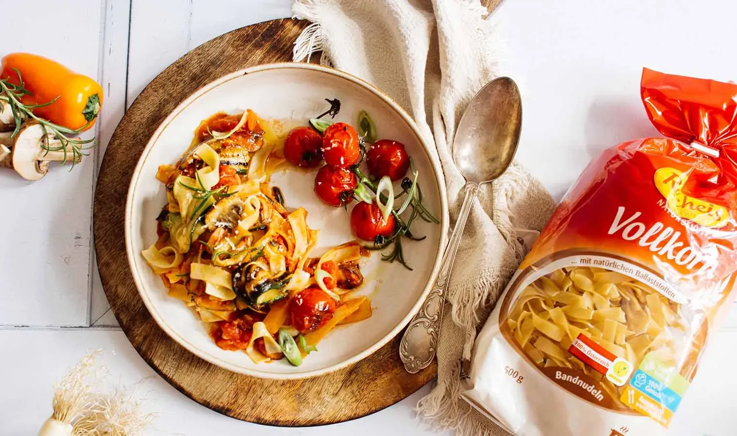 Ratatouille aus Grillgemüse-Resten mit Bandnudeln und Tomaten auf einem Teller neben Packung Vollkornnudeln und Löffel