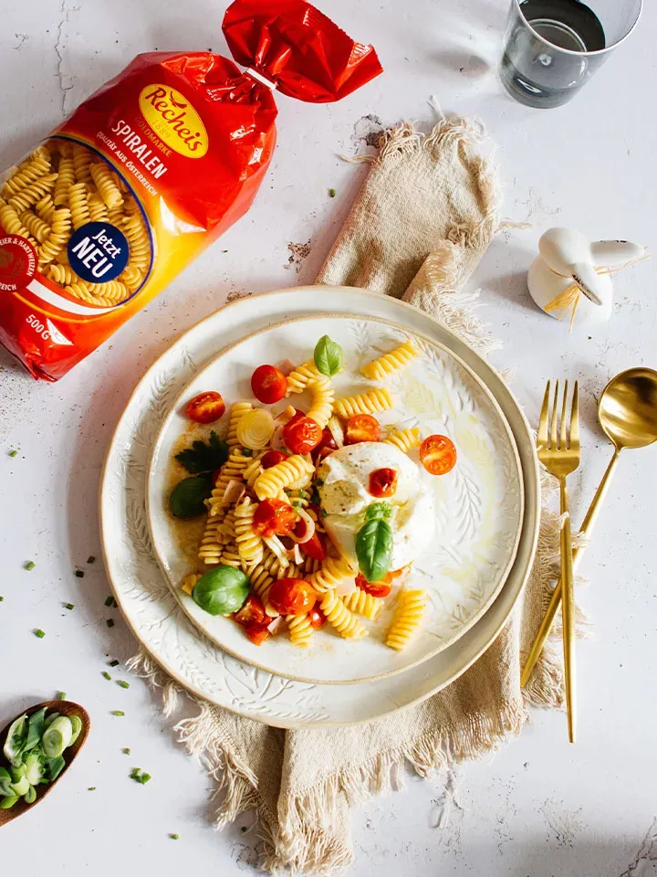 Restlrezept mit Osterschinken: Spiralnudeln, Tomaten und Mozzarella auf Teller neben Nudelpackung und Besteck