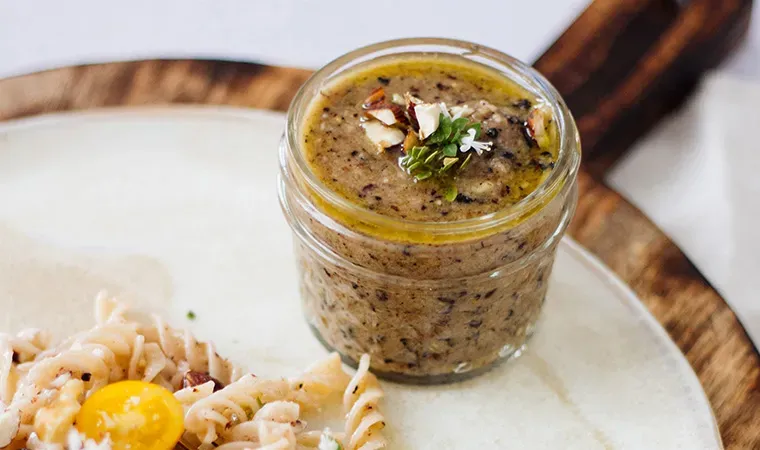 Glas mit braunem Aronia-Nuss-Pesto, mit Kräutern und Nüssen garniert, auf einem Teller