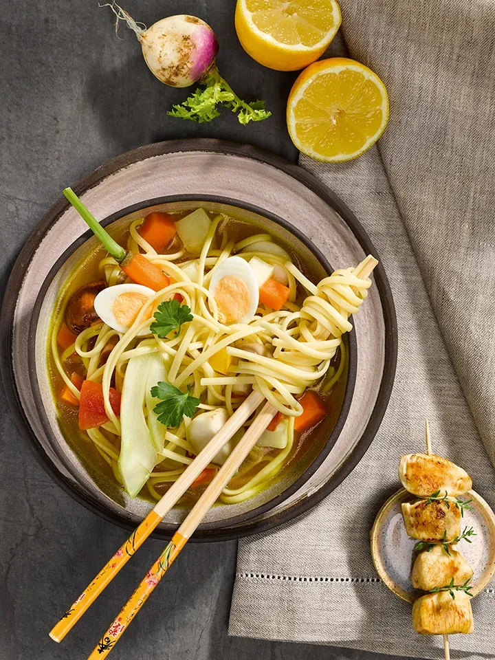 Asiatische Gemüsesuppe mit Linguine, Ei, Zitronenhälften und Teriyaki-Spießchen auf einer Stoffserviette