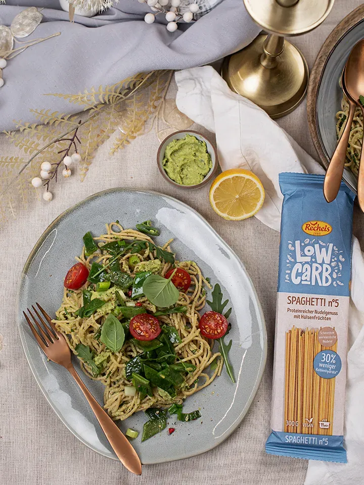 Low-Carb-Spaghetti mit Avocadopesto, Tomaten und grünem Gemüse auf einem Teller neben Pasta-Verpackung und Zitronenhälfte