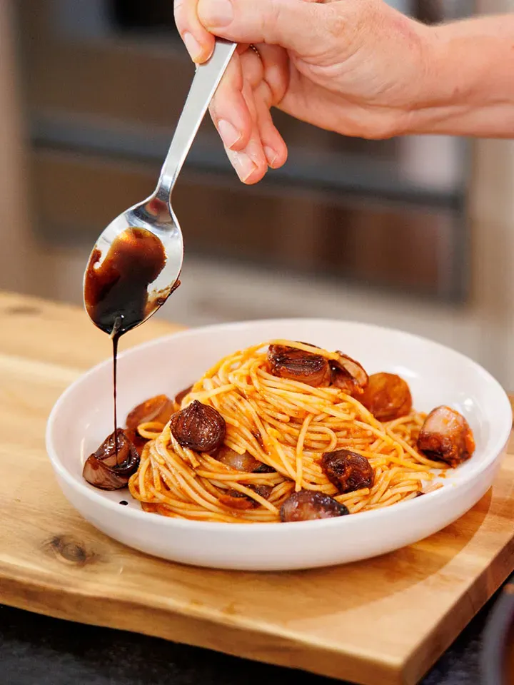 Hand träufelt dunkle Balsamico-Sauce auf Spaghetti mit gebratenem Knoblauch und Schalotten in einem weißen Teller