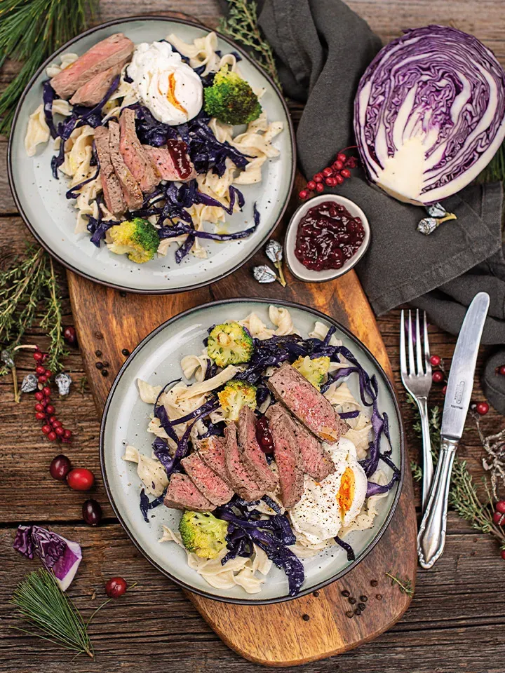 Bandnudeln mit Rinderfiletstreifen, Brokkoli, Rotkohl und einem Klecks Sauce auf zwei Tellern auf Holzbrett