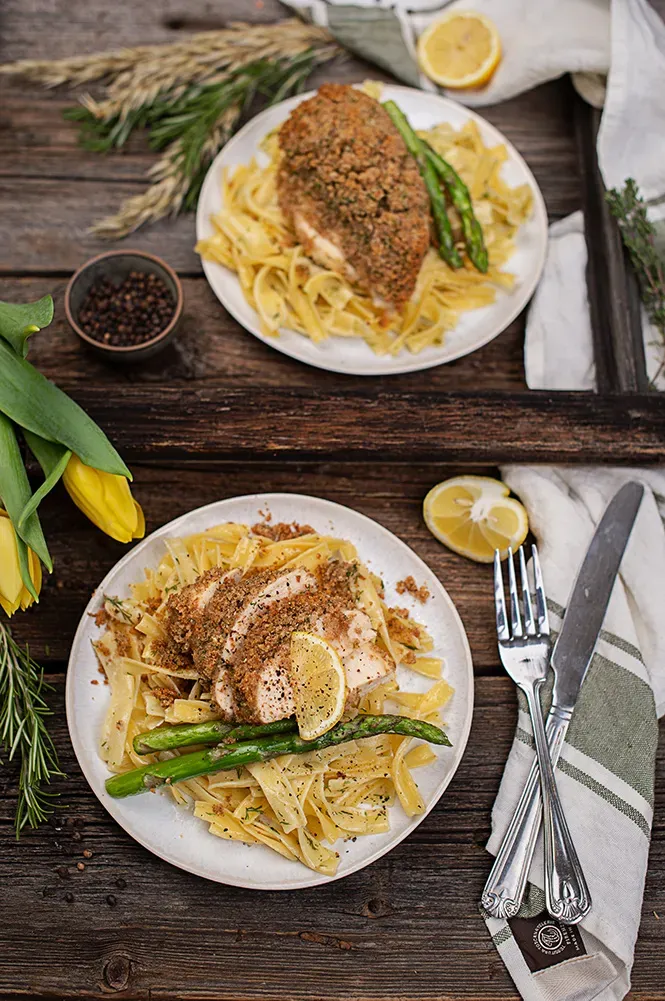 Bio-Bandnudeln mit Hühnerfilet in Parmesankruste, grünem Spargel und Zitronenscheiben auf zwei Tellern auf Holztisch